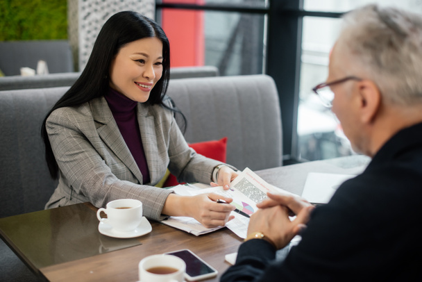 enfoque selectivo de sonriente mujer de negocios asiática hablando con hombre de negocios y apuntando a los papeles en la cafetería
  - Foto, Imagen