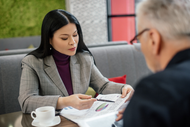 wybiórcze skupienie azjatyckiej bizneswoman rozmawiając z biznesmenem i wskazując na papiery w kawiarni  - Zdjęcie, obraz