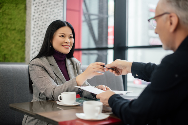 вибірковий фокус усміхненої азіатської бізнес-леді бере ручку від бізнесмена в кафе
  - Фото, зображення