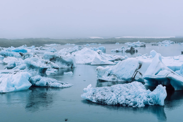 Heldere witte gletsjers bedekt met dichte mist in het blauwe water van het Jokulsalron gletsjermeer in IJsland - Foto, afbeelding