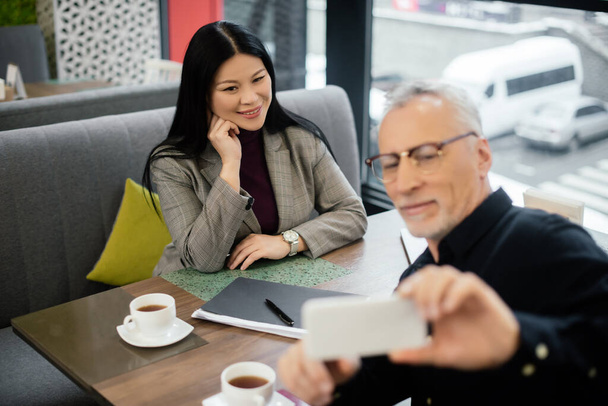 вибірковий фокус бізнесмена, беручи селфі з усміхненою азіатською бізнес-леді в кафе
  - Фото, зображення