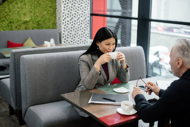 selektywne skupienie się uśmiechniętej azjatyckiej bizneswoman rozmawiając z biznesmenem i trzymając kubek w kawiarni  - Zdjęcie, obraz
