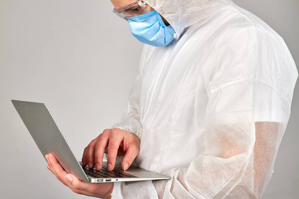 Beyaz koruyucu giysili adam dizüstü bilgisayar kullanarak solunum maskesi takıyor.. - Fotoğraf, Görsel