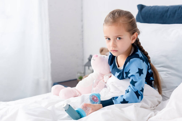 sick kid holding inhaler with spacer and looking at camera in bedroom  - Photo, Image