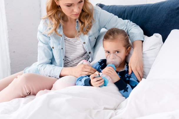 caring mother sitting near sick daughter using inhaler with spacer  - Фото, зображення