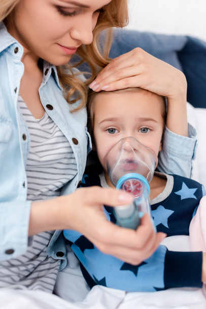 selective focus of attractive mother holding inhaler with spacer near sick daughter  - Fotografie, Obrázek