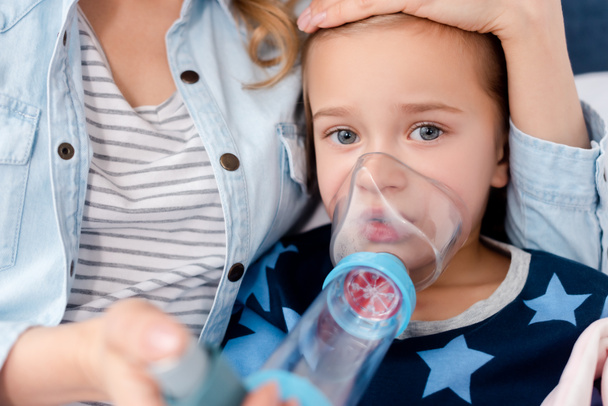 selective focus of caring mother holding inhaler with spacer near sick daughter  - Foto, immagini