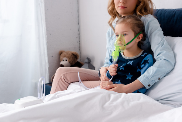mother sitting near sick daughter in respiratory mask using compressor inhaler  - Foto, Bild