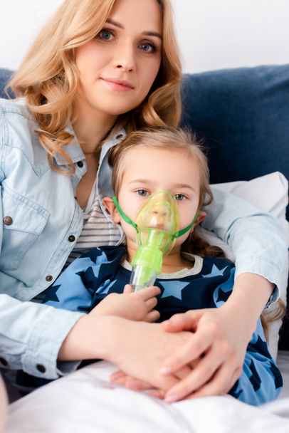 asthmatic kid in respiratory mask looking at camera near beautiful mother  - Фото, зображення