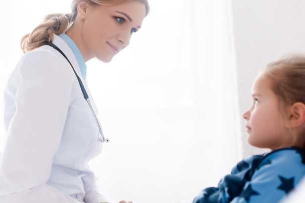 selective focus of attractive doctor in white coat looking at sick kid  - Фото, изображение