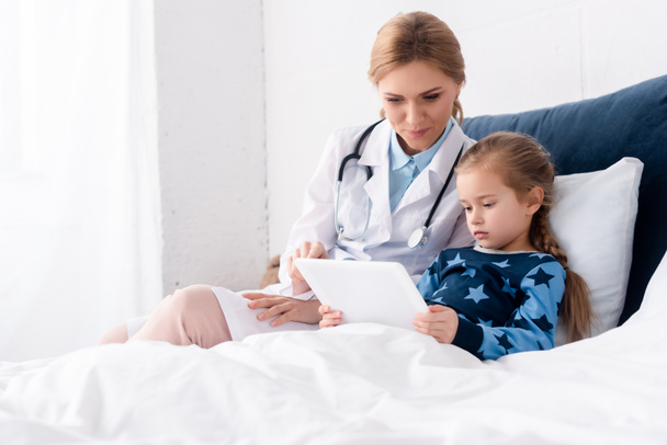 デジタルタブレットで病気の子供の近くに座っている白いコートの魅力的な医者  - 写真・画像