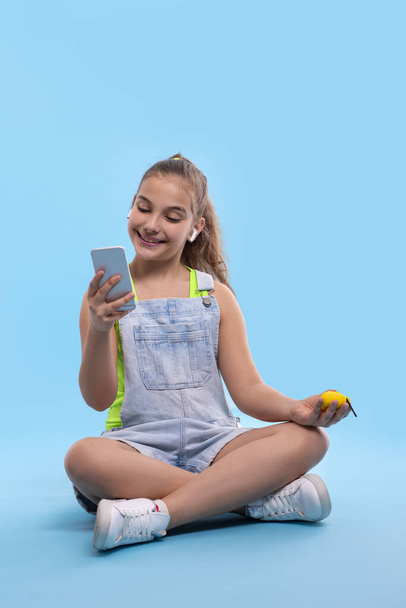 Ammuttu nuori brunette hymyilevä tyttö teini yllään denim haalarit shortsit istuu lootus asennossa ja katselee puhelimeen. Loma- ja reaktiokäsite
 - Valokuva, kuva