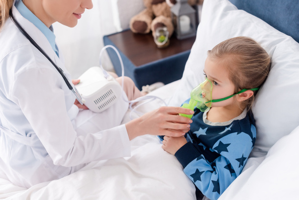 γιατρός σε λευκό παλτό αγγίζοντας αναπνευστική μάσκα σε ασθματικό παιδί χρησιμοποιώντας εισπνευστήρα συμπιεστή  - Φωτογραφία, εικόνα