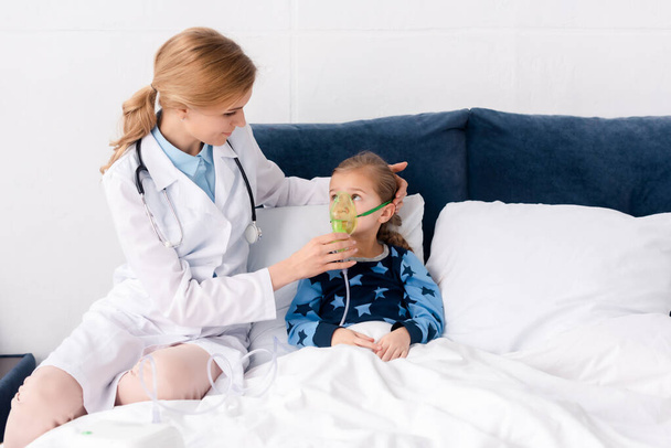όμορφη γιατρός σε λευκό παλτό αγγίζοντας αναπνευστική μάσκα σε ασθματικό παιδί  - Φωτογραφία, εικόνα