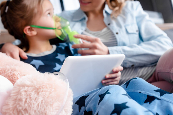 selektiver Fokus asthmatischer Kinder mit Atemmaske und digitalem Tablet in der Nähe von Mutter und Plüschtier  - Foto, Bild