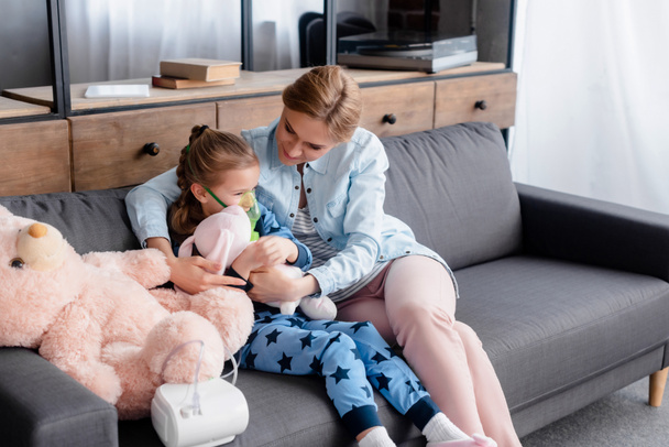 niño asmático usando inhalador de compresor cerca de madre cariñosa con juguete suave
  - Foto, imagen