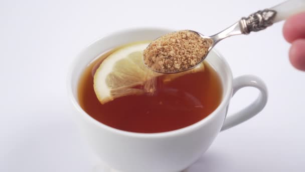 mano aggiunge zucchero di canna con un cucchiaio da dessert in una tazza con tè nero caldo e con una fetta di limone primo piano. bevanda vitaminica calda per migliorare l'immunità
 - Filmati, video