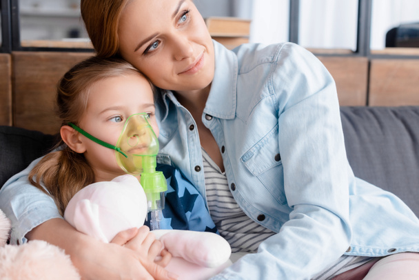 mother hugging asthmatic daughter in respiratory mask - Фото, изображение