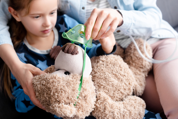 selective focus of mother touching respiratory mask on teddy bear near sick kid  - Photo, Image