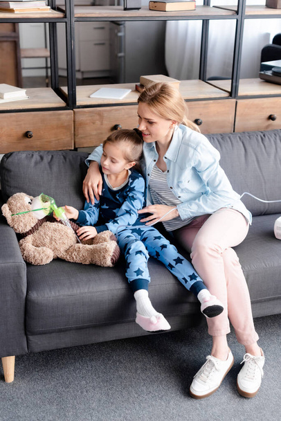 Kind berührt Atemmaske auf Teddybär neben Mutter auf Sofa - Foto, Bild