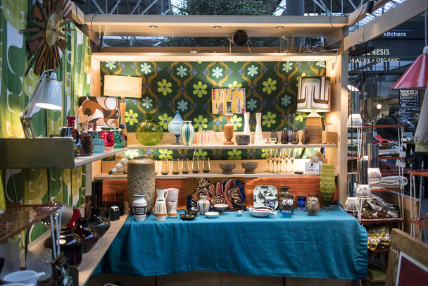 London, UK - 13 February, 2020 Spitalfields flea market. Glass vases and chandeliers in the style of the seventies on the shelves on the background of multi-colored fabric in a green flower - Photo, Image