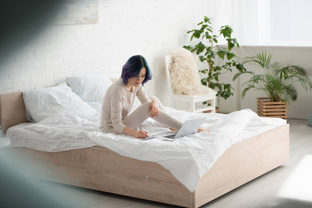 Selectieve focus van freelancer met kleurrijk haar schrijven in notebook in de buurt laptop op bed in de slaapkamer - Foto, afbeelding