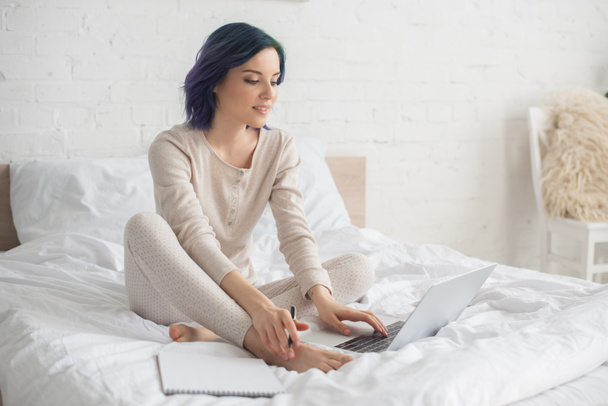 Freelancer with colorful hair holding pen and working on laptop near copybook on bed - Foto, Imagem