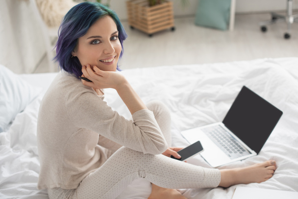Vista de alto ângulo do freelancer com cabelo colorido, caneta e smartphone sorrindo e olhando para a câmera perto do laptop na cama
 - Foto, Imagem