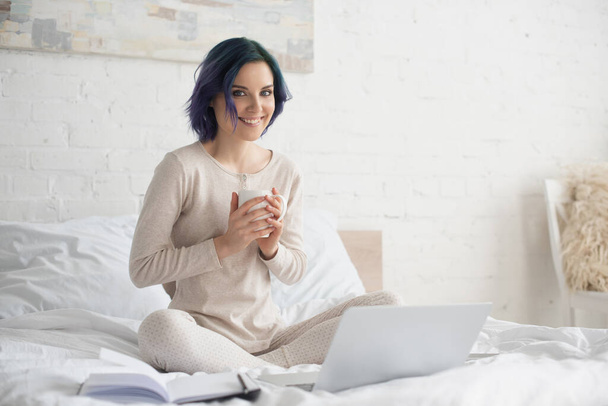 Freelancer met kleurrijk haar en gekruiste benen houden kopje thee en kijken naar camera in de buurt van laptop en notebook op bed - Foto, afbeelding
