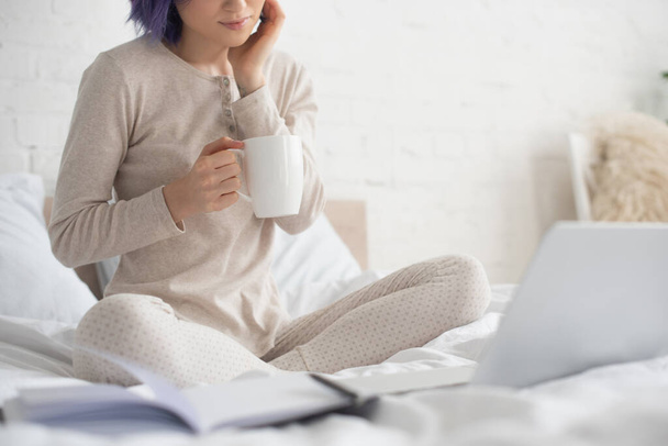 Vista recortada de freelancer con piernas cruzadas sosteniendo taza de té cerca de la computadora portátil en la cama
 - Foto, imagen