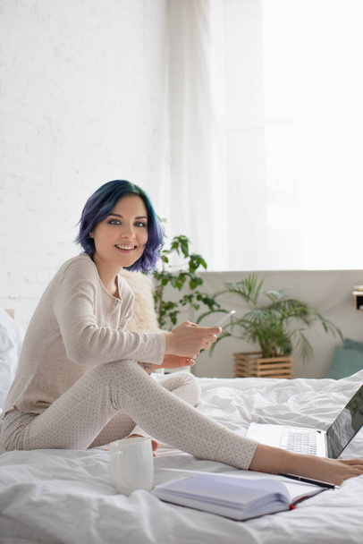 Freelancer with colorful hair smiling, looking at camera and using smartphone near laptop, notebook and cup of tea on bed - Fotografie, Obrázek