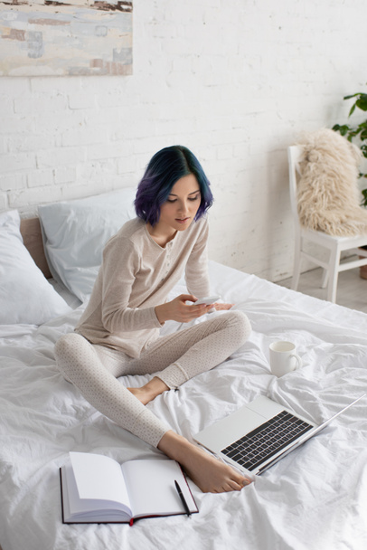 Высокоугольный вид красивого фрилансера с раскрашенными волосами с помощью смартфона возле ноутбука и ноутбука на кровати в спальне
 - Фото, изображение