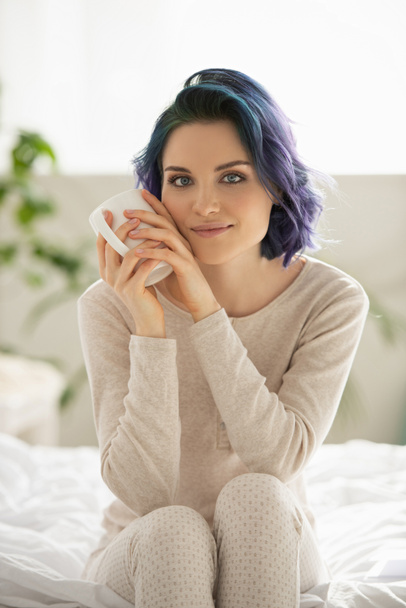 Beautiful girl with colorful hair holding cup of tea, looking at camera and smiling on bed in bedroom - Фото, зображення