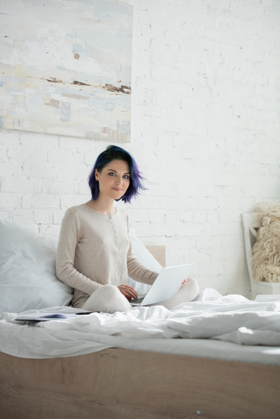 Freelancer with colorful hair and crossed legs working with laptop, smiling and looking at camera on bed in bedroom - 写真・画像
