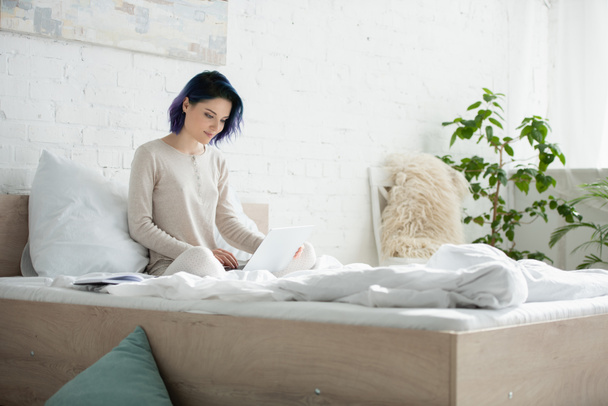 Freelancer with colorful hair and crossed legs working with laptop and smiling on bed in bedroom - Foto, afbeelding