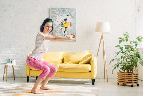 Girl with colorful hair in chair pose with outstretched hands looking at camera in living room - Photo, Image