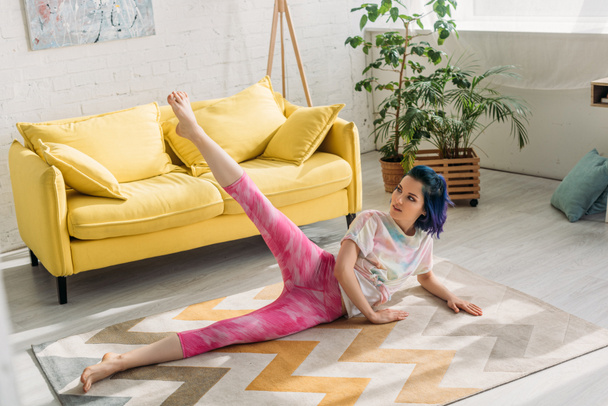 Vista de ángulo alto de la chica con el pelo colorido estiramiento en el suelo en la sala de estar
 - Foto, Imagen