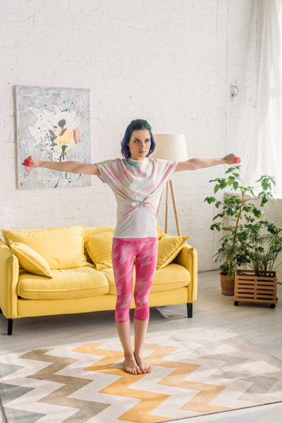 Tense woman with colorful hair holding dumbbells with outstretched hands in living room - Foto, Imagen