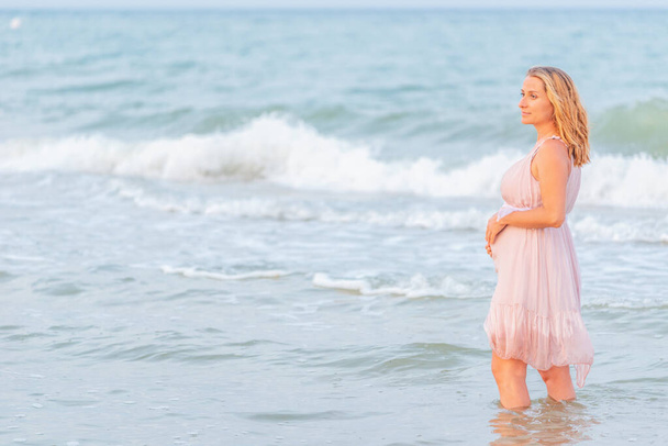 Beautiful happy young pregnant woman in a summer light dress hugging her belly and walks along the sea waves on a sunny summer evening during vacation. Natural Parenthood Concept. Copyspace - Φωτογραφία, εικόνα