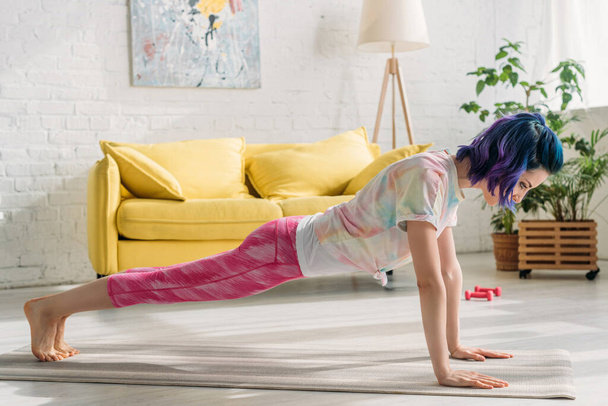 Woman with colorful hair doing plank on yoga mat in living room - Zdjęcie, obraz