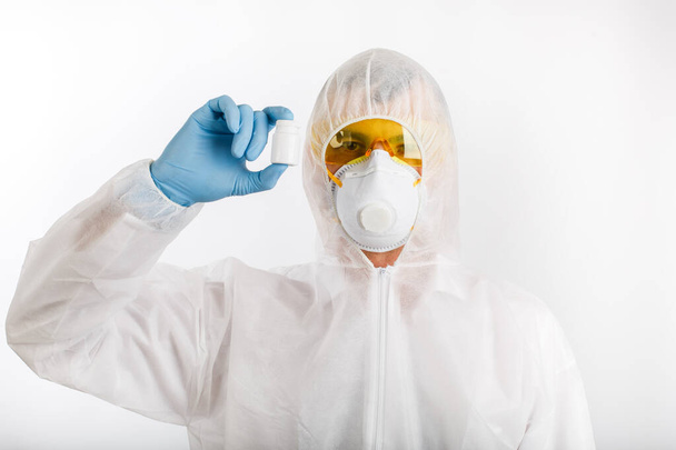 Doctor in disposable anti-epidemic suit wearing a respirator and glasses holds bottle with pills on white background - Fotografie, Obrázek