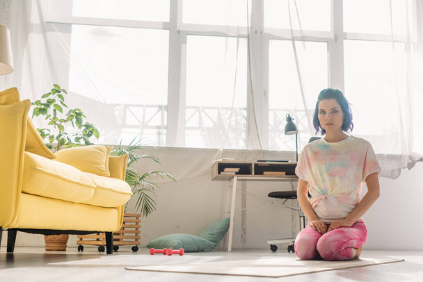 Dívka s barevnými vlasy dívá do kamery a sedí na podložce jógy v obývacím pokoji - Fotografie, Obrázek