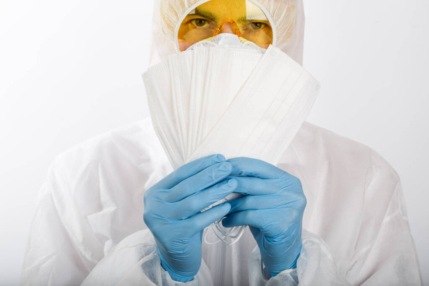 Доктор в одноразовому анти-епідемічному костюмі в респіраторі і окулярах тримає медичні маски для захисту від вірусу інфекції на білому тлі
 - Фото, зображення