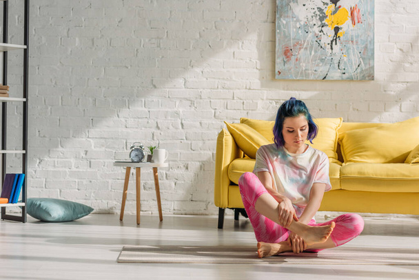 meisje met kleurrijk haar aanraken been op yoga mat in woonkamer - Foto, afbeelding