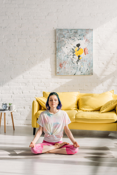 Mädchen mit buntem Haar und geschlossenen Augen meditiert auf Yogamatte im Wohnzimmer - Foto, Bild