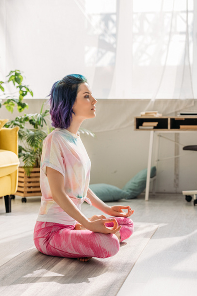 Frau mit buntem Haar meditiert auf Yogamatte im Wohnzimmer - Foto, Bild