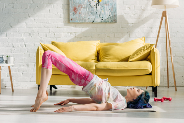 Fille aux cheveux colorés en pose de pont sur tapis de yoga dans le salon
 - Photo, image