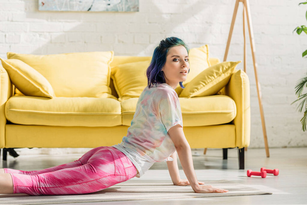 Chica con el pelo colorido en cobra pose en estera de yoga mirando a la cámara en la sala de estar
 - Foto, Imagen