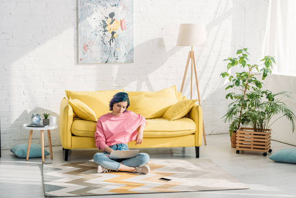 Freelancer with colorful hair and crossed legs working with laptop near sofa on floor  - Photo, Image