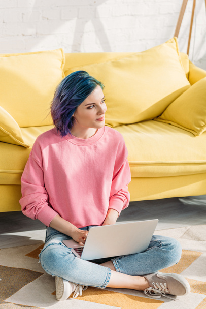 Thoughtful freelancer with colorful hair looking away and working with laptop near sofa on floor in living room - Photo, image